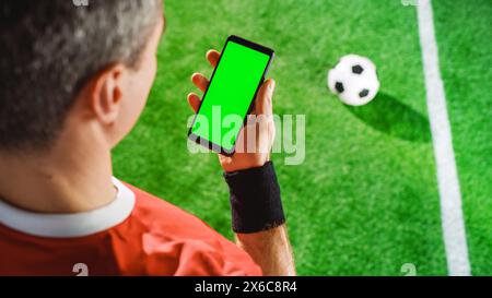 Profi-Fußballspieler mit Smartphone mit grünem Bildschirm Chroma Key mit Tracking Markern. Erhöhte Top Down Over Shoulder Aufnahme eines Fußballstars im Internet, während er auf dem Spielfeld steht Stockfoto