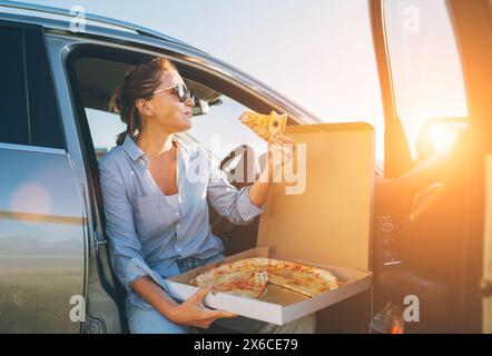 Eine Frau mittleren Alters isst gerade italienische Pizza, sitzt auf dem Fahrersitz während der Essenspause und genießt den Sonnenuntergang. Autofahrt, Fast Food je Stockfoto