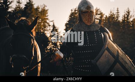 Der mittelalterliche Ritterkrieger führt sein Pferd in die Schlacht, um zu kämpfen. Porträt des Helden in überzogener Rüstung, Schild, Helm, Schwert und Vollbluthengst. Filmische Licht, Krieg, Rauch, Invasion, Eroberung Stockfoto