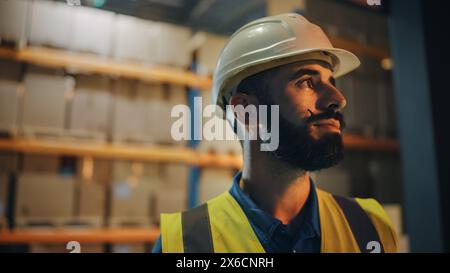 Porträt eines männlichen lateinischen Arbeiters, der einen harten Hut trägt und vor dem Lager lächelnd steht. Professioneller Frontline-Held mit stilvollem Bart im Kinolicht Stockfoto