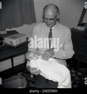 1960er Jahre, historisch, ein älterer Herr, ein Großvater, der auf einem Stuhl in einem Vorderzimmer sitzt, mit einem Baby auf dem Schoß, das es mit einer kleinen Flasche füttert. Neben ihm auf einem kleinen Tisch, einem 35-mm-Diaprojektor. Stockfoto