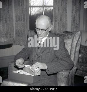 1960er Jahre, historisch, ein älterer Herr in Anzug und Krawatte, der in einem Klubsessel sitzt und 35-mm-Filmdias in einem Tablett ansieht. Fotografische Dias stammen aus dem Jahr 1935, als Kodak Kodachome Diafilm einführte, der schnell das beliebteste Filmformat aller Zeiten einführte. 35-mm-Folien werden auch als Fotofolien bezeichnet, da sie über ein Fenster verfügen, in dem der Zuschauer den physischen Film sehen kann. Stockfoto