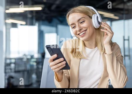 Glückliche junge Frau, die Kopfhörer verwendet und gleichzeitig ein Smartphone in einem stilvollen Büro hält, um einen Moment der Freude und Entspannung bei der Arbeit zu zeigen. Stockfoto