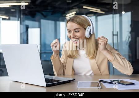Die fröhliche blonde Frau mit Kopfhörern feiert einen Sieg, während sie auf ihrem Laptop in einer modernen Büroumgebung ein Sportereignis ansieht. Stockfoto