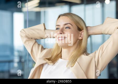 Eine professionelle blonde Frau in einem beigefarbenen Blazer macht eine entspannende Pause in ihrem Büro, lächelt zufrieden und lehnt sich mit den Händen hinter dem Kopf zurück. Stockfoto