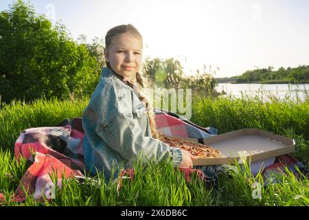 Ein Mädchen in einem blauen Denim-Anzug sitzt auf einer Decke auf dem grünen Gras neben einer Pizzaschachtel. Sonniger Sommertag am Flussufer. Picknick im Freien Stockfoto