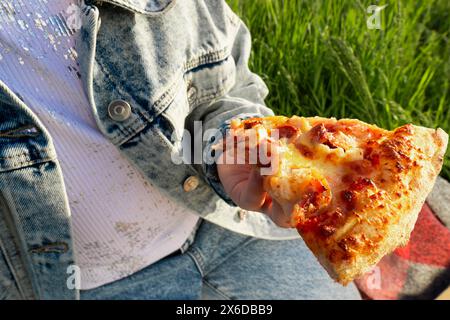 Ein Stück Pizza in den Händen vor dem Hintergrund einer Decke auf grünem Gras. Picknick im Freien. Nahaufnahme Stockfoto