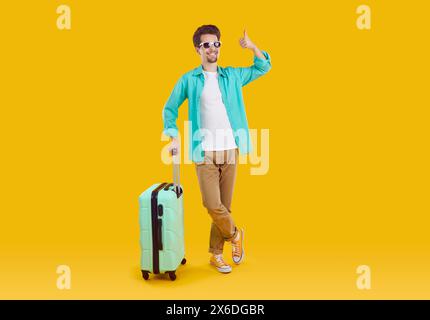 Glücklicher, fröhlicher Mann, der billige Flugtickets gekauft hat, zeigt Daumen hoch, während er mit Reisekoffer posiert. Stockfoto