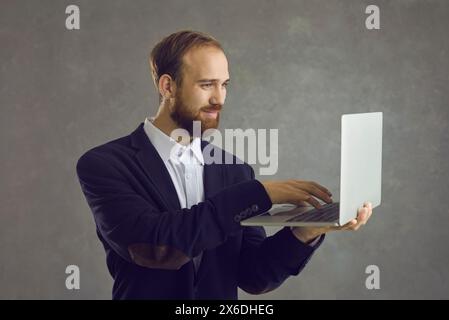 Der europäische Mann steht auf einem grauen Hintergrund und schreibt Text auf einen Laptop, den er in der Hand hält. Stockfoto