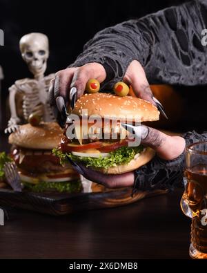 Die Hexenhände halten einen Monster Burger in ihren Händen auf einem sitzenden Skelett. Die perfekte Vorspeise für die Halloween Party Stockfoto