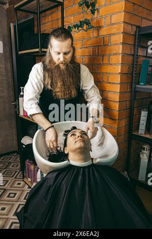 Der Friseur wäscht die Haare der Frau. Mann junger Friseur-Profi, der Erfahrung in Weste mit langem Bart und Haar hat, Haarwäschekopf weibliche Klientin in Stockfoto