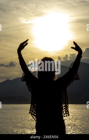 Dieses ergreifende Bild fängt die Silhouette einer wunderschönen Frau ein, deren Arme in einer anmutigen Umarmung der verblassenden Sonne hochgezogen sind. Stockfoto