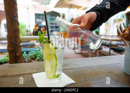 Limonade mit frischer Zitrone auf Holztischhintergrund. Die Hand eines Mannes in einem schwarzen Ärmel gießt ein kohlensäurehaltiges Getränk in ein hohes, durchsichtiges Glas, in dem Stockfoto