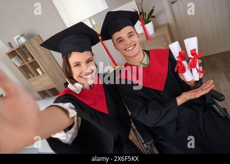 Männlicher Rollstuhlabsolvent mit Diplomen und sein Klassenkamerad macht zu Hause Selfie Stockfoto