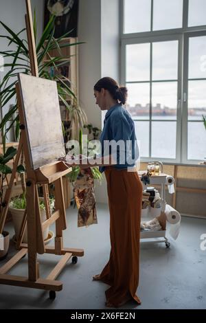 Friedliche Künstlerin mit Pinsel arbeitet kleine Details von kleinen Gemälden auf Staffelei aus. Stockfoto