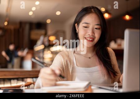 Eine junge positive, glückliche asiatische Studentin, die online studiert und Hausaufgaben in einem Coffee Shop in der Stadt macht, macht Notizen in ihrem Notizbuch, Papa Stockfoto