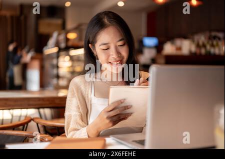 Eine junge positive, glückliche asiatische Studentin, die online studiert und Hausaufgaben in einem Coffee Shop in der Stadt macht, macht Notizen in ihrem Notizbuch, Papa Stockfoto