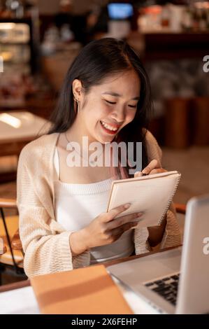 Eine junge positive, glückliche asiatische Studentin, die online studiert und Hausaufgaben in einem Coffee Shop in der Stadt macht, macht Notizen in ihrem Notizbuch, Papa Stockfoto