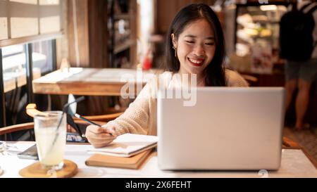 Eine junge positive, glückliche asiatische Studentin, die online studiert und Hausaufgaben in einem Coffee Shop in der Stadt macht, macht Notizen in ihrem Notizbuch, Papa Stockfoto