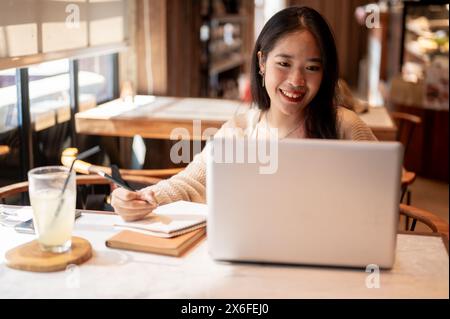 Eine junge positive, glückliche asiatische Studentin, die online studiert und Hausaufgaben in einem Coffee Shop in der Stadt macht, macht Notizen in ihrem Notizbuch, Papa Stockfoto