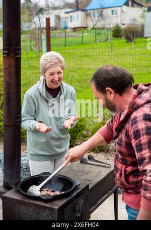 Reifer russischer Mann und Frau, die Fleisch in einem Kessel auf dem Herd im Freien braten, Familienkonzept Stockfoto