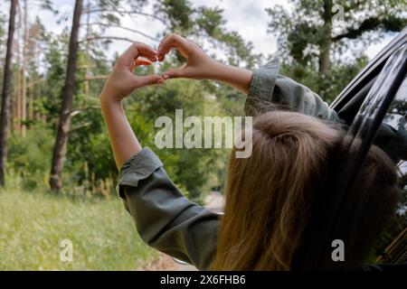 Frau, die im Auto fährt, die Hand aus dem Fenster hält und ein Herzsymbol mit ihren Händen zeigt. Love and Adventure Love Love and Adventure Local Travel Freiheit und Verbindung mit der Natur. Roadtrip mit nachhaltigem Lebensstil am Wochenende zum Entspannen und Ausruhen Stockfoto