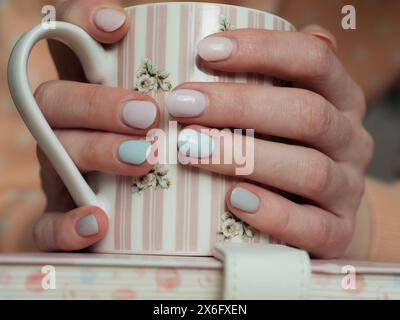 Horizontale Nahaufnahme von Frauenhänden, die einen niedlichen Becher auf einem Notizbuch umarmen – alles in Pastellfarben in Blau, Pfirsichfarben und Rosa. Stockfoto