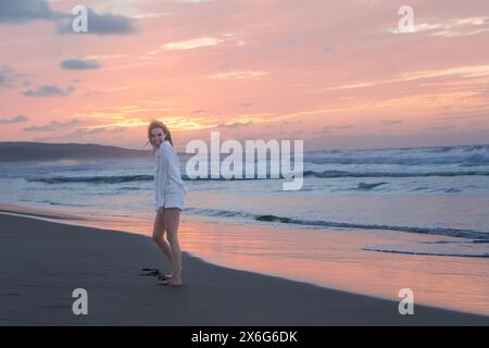 Die junge Frau genießt einen friedlichen Spaziergang entlang der Küste, während die Sonne untergeht und ein sanftes Leuchten über den Sandstrand und die sanften Meereswellen wirft Stockfoto