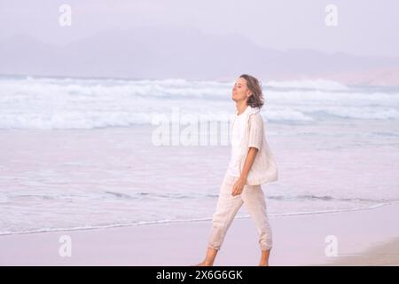 Junge Frau genießt einen friedlichen Spaziergang entlang eines Sandstrandes mit Wellen, die sanft pastellfarbene Sonnenuntergänge schaffen eine ruhige Kulisse Stockfoto