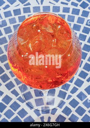 Nahaufnahme eines Aperol Spritz Cocktails mit Eiswürfeln, im Sonnenlicht glühend, auf einer gemusterten Oberfläche Stockfoto