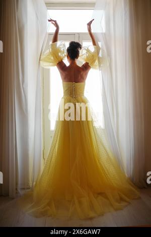 Die Silhouette einer Frau in einem goldenen, luxuriösen Kleid vor dem Hintergrund eines Fensters hält einen Vorhang mit den Händen. Elegante Dame in gelber Seide Stockfoto