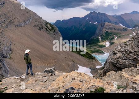 Gereifter kaukasischer Mann, der am Rande einer Klippe steht und die Szene eines Gletschersees in einer tiefen Schlucht mit Schneeflecken und hohen Bergen beobachtet Stockfoto