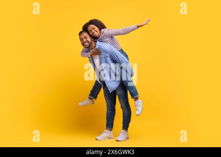 Fröhliches Paar genießt einen Huckepack-Ritt vor einem hellen gelben Hintergrund Stockfoto