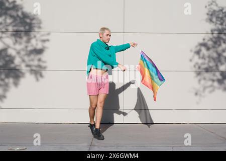 Junger Lateinmann mit kurzen blonden Haaren, rosa Shorts, schwarzen Turnschuhen und grünem Pullover, ist draußen mit Stolz-Flagge, die tanzt und den Sonnenuntergang genießt Stockfoto