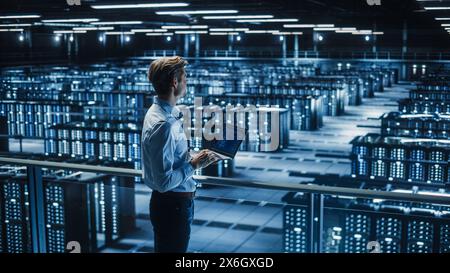 Hübscher, lächelnder IT-Spezialist mit Tablet-Computer im Rechenzentrum. Erfolgreicher Geschäftsmann und E-Business-Unternehmer mit Blick auf die Big Server Farm für Cloud Computing. Stockfoto