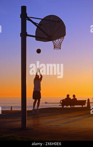 Ein erwachsener Mann übt seine Basketball-Fähigkeiten in einem Park nahe der Küste in Laguna Beach, Kalifornien, während der Sonnenuntergang ihn in Silhouette verwandelt Stockfoto