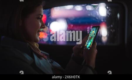 Happy Female pendelt nachts auf dem Rücksitz eines Taxis nach Hause. Schöne Passagierin, die Arcade-Videospiel auf Smartphone spielt, während sie in einem Auto in der Urban City Street mit funktionierenden Neonschildern sitzt. Stockfoto