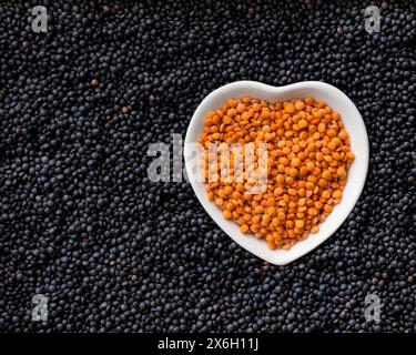 Im Vordergrund eine herzförmige weiße Schüssel mit getrockneten roten Linsen auf einem Hintergrund aus schwarzen Linsen. Stockfoto