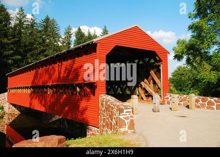 Eine kleine Landstraße überquert einen Bach über eine malerische und charmante überdachte Brücke aus Holz Stockfoto