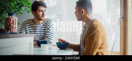 Zwei gutaussehende Freunde essen buntes Frühstückszerealien in der gemütlichen Küche im stilvollen Apartment. Junge Erwachsene Schwule Paare haben ein Gespräch beim Essen gesunde nahrhafte Nahrung am Morgen. Stockfoto