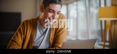 Hübscher Erwachsener mit Laptop-Computer, im Wohnzimmer in der Wohnung zu Hause sitzend. Attraktiver Mann ist Online-Shopping im Internet, erledigt Fernarbeit oder schaut lustige Videos im Streaming-Service an. Stockfoto