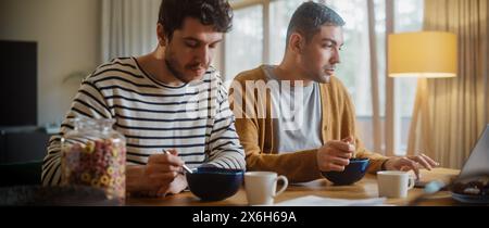 Zwei gutaussehende Freunde essen buntes Frühstückszerealien in der gemütlichen Küche im stilvollen Apartment. Junge Erwachsene Schwule Paare haben ein Gespräch beim Essen gesunde nahrhafte Nahrung am Morgen. Stockfoto
