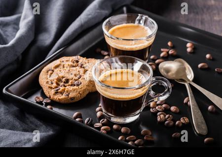 Auf dem schwarzen Tablett, im Vordergrund, zwei duftende Kaffees in Glasbechern und Schokoladenkekse Stockfoto