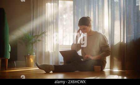 Schöne glückliche Frau mit Laptop zu Hause für Fernarbeit. Junge Erwachsene lächelnde Frau, die auf dem Boden sitzt, nutzt das Internet, schreibt Bericht, beendet Projekt. Guten sonnigen Tag für produktive Fernarbeit Stockfoto