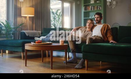 Paare Watches Fernsehen, während sie auf einer Couch im Wohnzimmer sitzen. Freundin und Freund lügen umarmt. Verbringen Sie ein Wochenende zusammen zu Hause mit stilvollem Interieur. Stockfoto