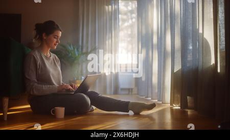 Schöne glückliche Frau mit Laptop zu Hause für Fernarbeit. Junge Erwachsene lächelnde Frau, die auf dem Boden sitzt, nutzt das Internet, schreibt Bericht, beendet Projekt. Guten sonnigen Tag für produktive Fernarbeit Stockfoto