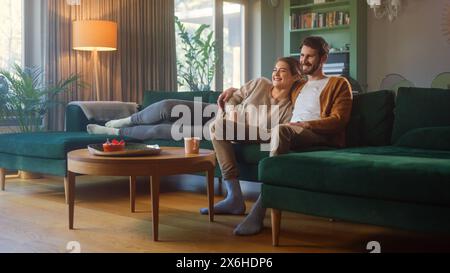 Paare Watches zusammen, während du auf einer Couch im Wohnzimmer sitzt. Freundin und Freund umarmen, kuscheln, reden, lächeln und Fernsehen Streaming Services. Zuhause mit gemütlichem, stilvollem Interieur. Stockfoto