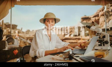 Archäologische Grabstätte: Berühmte Archäologin lächelt vor der Kamera und verwendet Laptop, analysiert fossile Überreste der antiken Zivilisation Kulturartefakte. Ein Team von Historikern arbeitet an der Ausgrabungsstätte Stockfoto