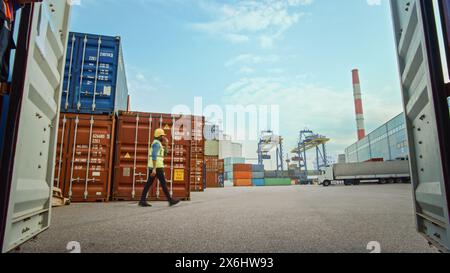 Gutaussehender afrikanisch-amerikanischer schwarzer Industrieingenieur in gelbem Schutzhelm und Sicherheitsweste, der bei einem Job im Containerterminal unterwegs ist. Foreman oder Supervisor betreiben die Logistik. Stockfoto