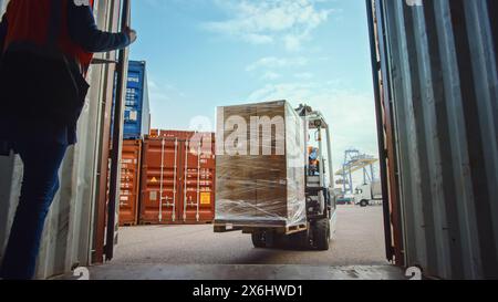 Gabelstaplerfahrer, der einen Frachtcontainer mit einer vollen Palette mit Kartonkisten im Logistics Operations Port Terminal beladen. Weibliche Betriebsleiterin und Sicherheitsinspektorin helfen dem Prozess. Stockfoto